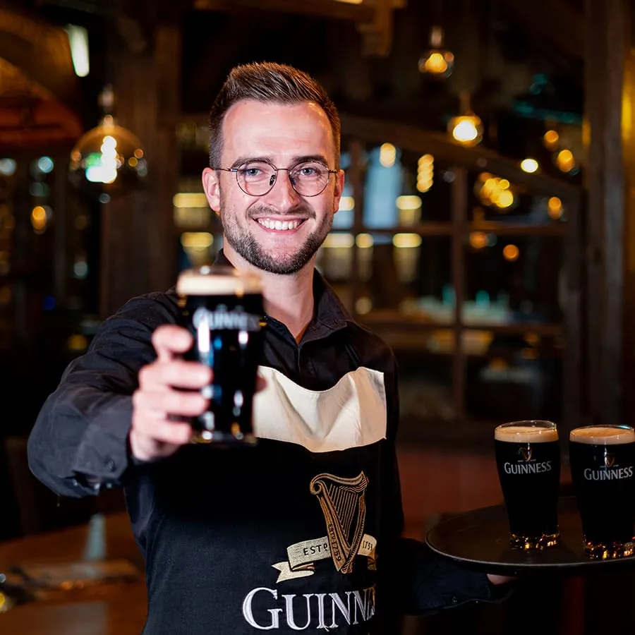 Ein Mitarbeiter mit Guinness-Schürze hält ein frisch gezapftes dunkles Bier in die Kamera
