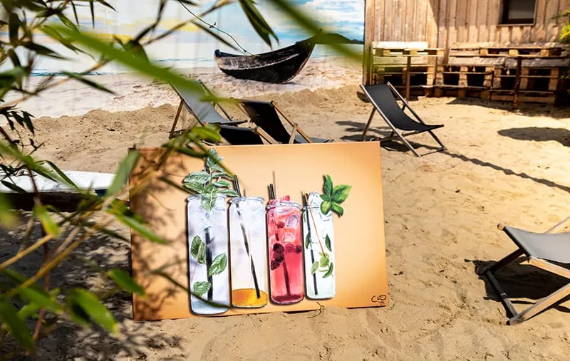 Gezeichnete Limonadengläser auf einem Bild, das am Strand im Sand steht, dahinter Liegestühle