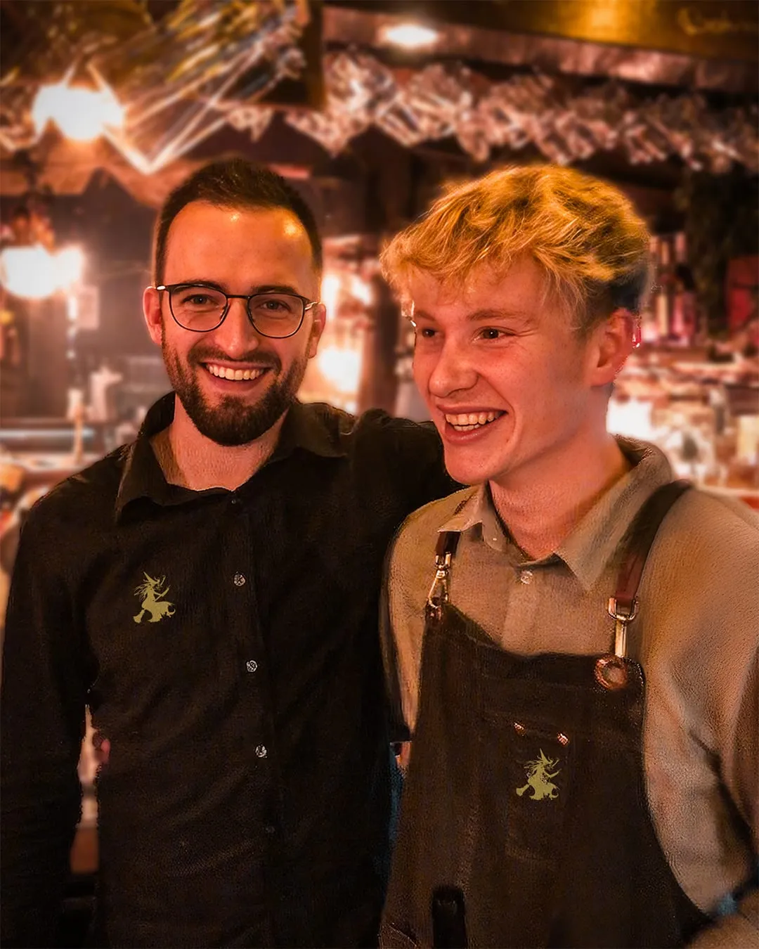 Zwei Mitarbeiter in dunklen Hemden und Schürze mit Hexen-Logo lachen im Gastraum in die Kamera